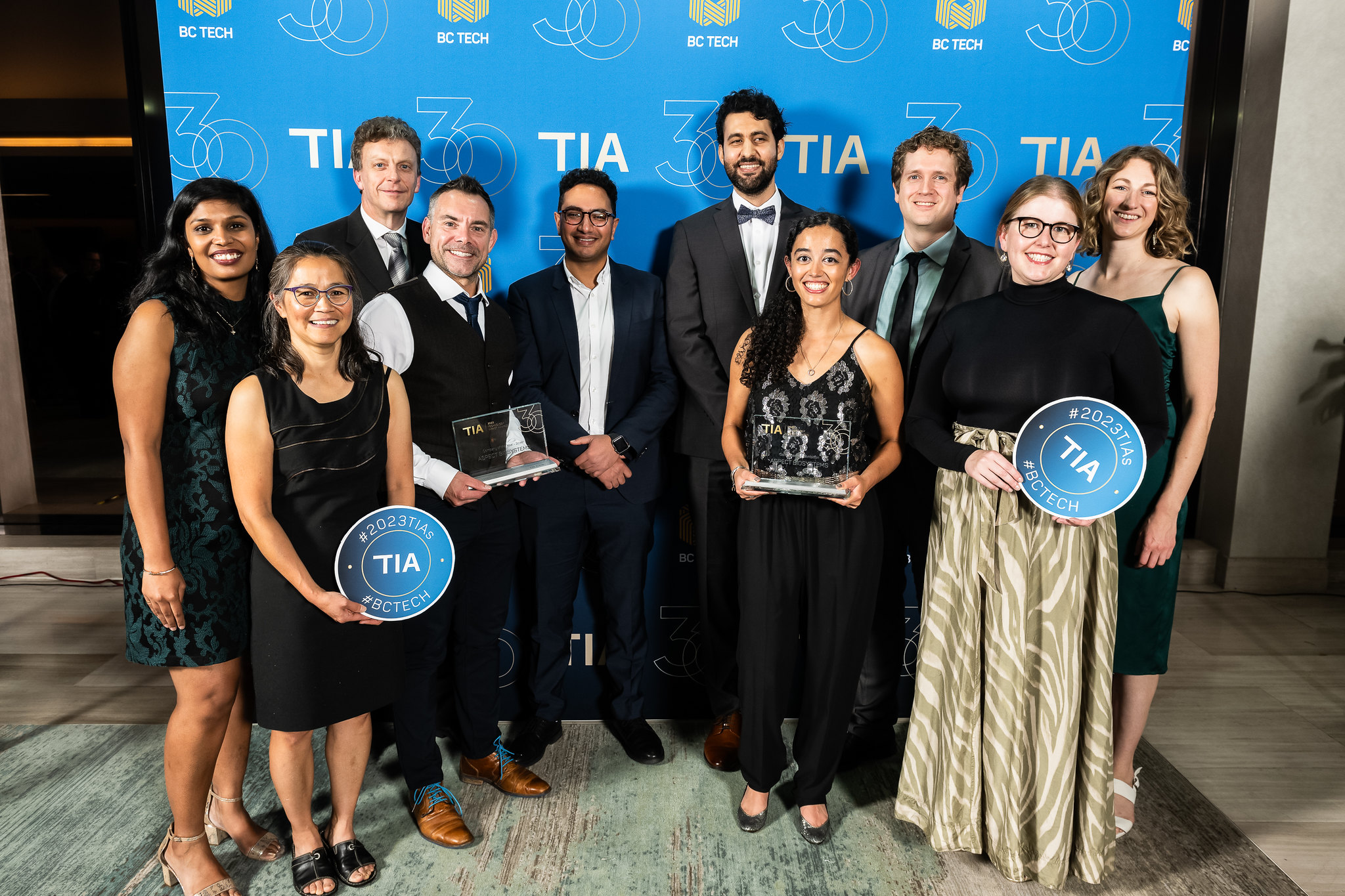 10 people from the Aspect Biosystem in formal attire, standing in front of a blue photo backdrop and holding two glass awards.